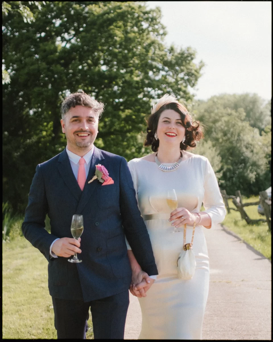 A smiling couple walks hand-in-hand outdoors at a Yoghurt Rooms wedding festival, dressed in formal attire. The man wears a navy suit with a pink tie; the woman, in an elegant white dress, holds a glass of champagne amid green trees and grass.