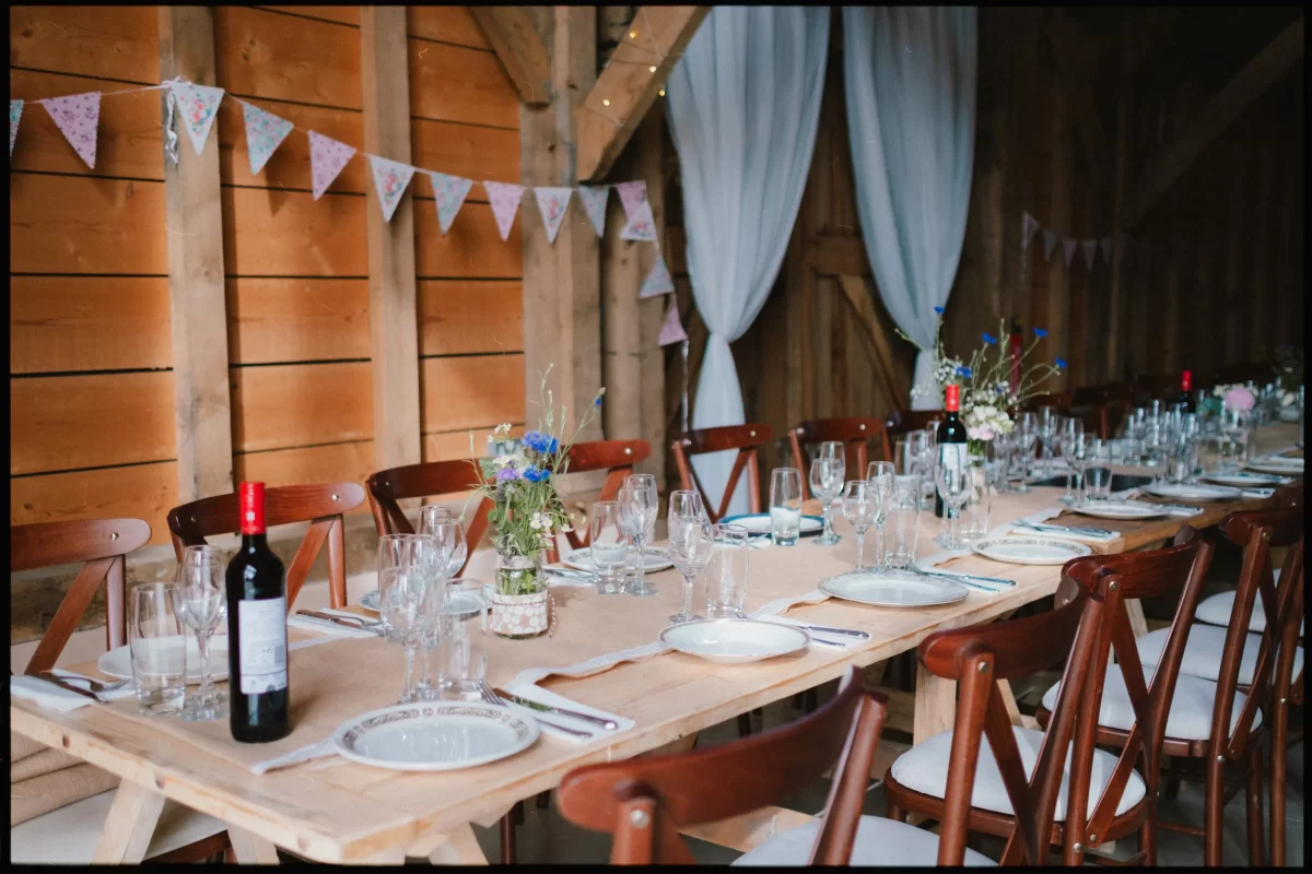 A rustic wooden table set for a meal with plates, cutlery, wine glasses, bottles of red wine, and small flower arrangements; bunting and white curtains decorate the barn-style interior, perfect for a Yoghurt Rooms wedding festival celebration.