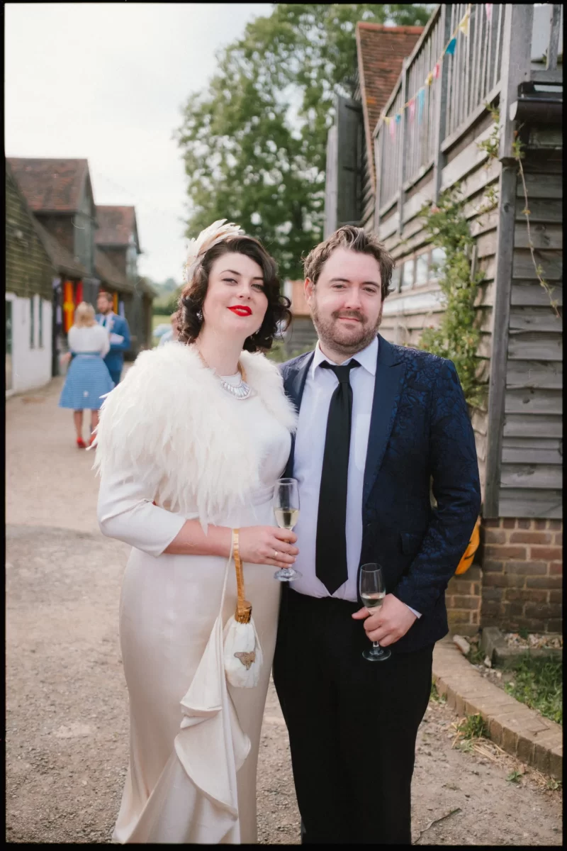 A woman in a vintage white dress and feathered shawl stands beside a man in a dark suit and tie. Both hold champagne glasses and smile outdoors at a Yoghurt Rooms wedding festival near rustic wooden buildings.