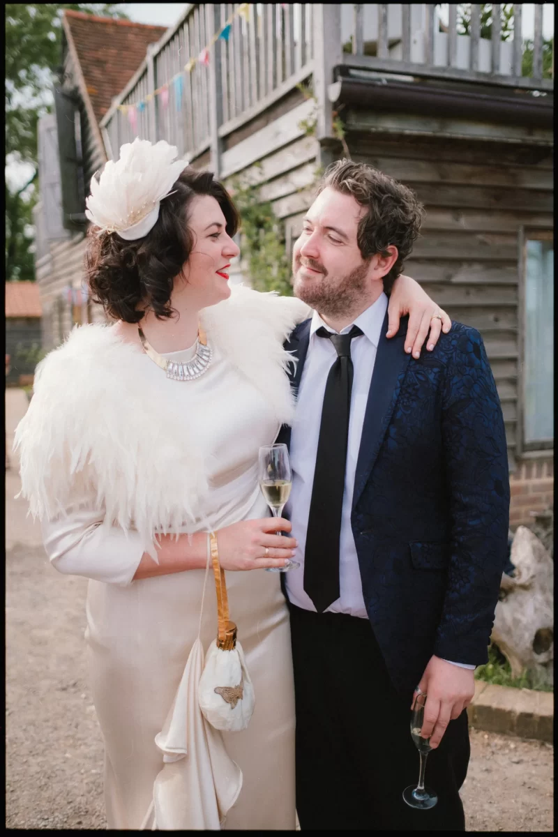 A woman in a white dress and feathered shawl smiles with her arm around a man in a blue suit and black tie. Both hold champagne glasses, standing outside by a rustic wooden building at a Yoghurt Rooms wedding festival.