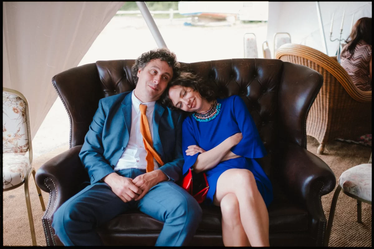 A man in a blue suit and orange tie sits on a dark leather sofa, leaning his head toward a woman in a blue dress who is resting her head on his shoulder with her eyes closed; they look relaxed and content after the lively yoghurt rooms wedding festival.