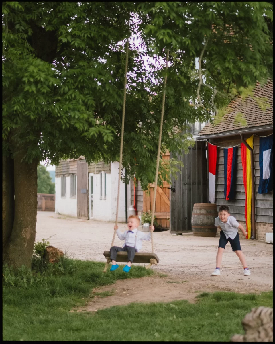 Two young children play outside near the yoghurt rooms wedding festival; one boy pushes another on a wooden swing by a rustic building adorned with colorful flags and a large wooden barrel in the background.