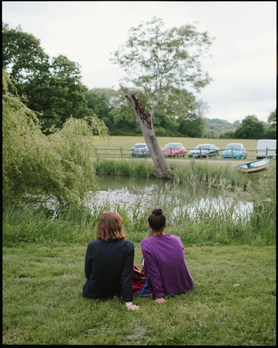 Two people sit on grass near a pond at a yoghurt rooms wedding festival, surrounded by trees, tall grasses, and parked cars across the water. The peaceful scene captures the beauty of nature in an outdoor setting.