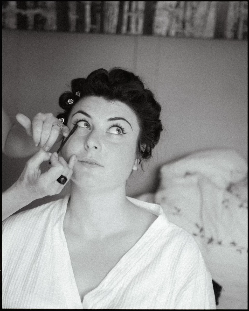A woman in a bathrobe with hair rollers looks upward as someone applies makeup to her lower eyelid, preparing for a wedding festival. The cozy indoor scene features soft light, a bed, and the calm ambiance of yoghurt rooms.