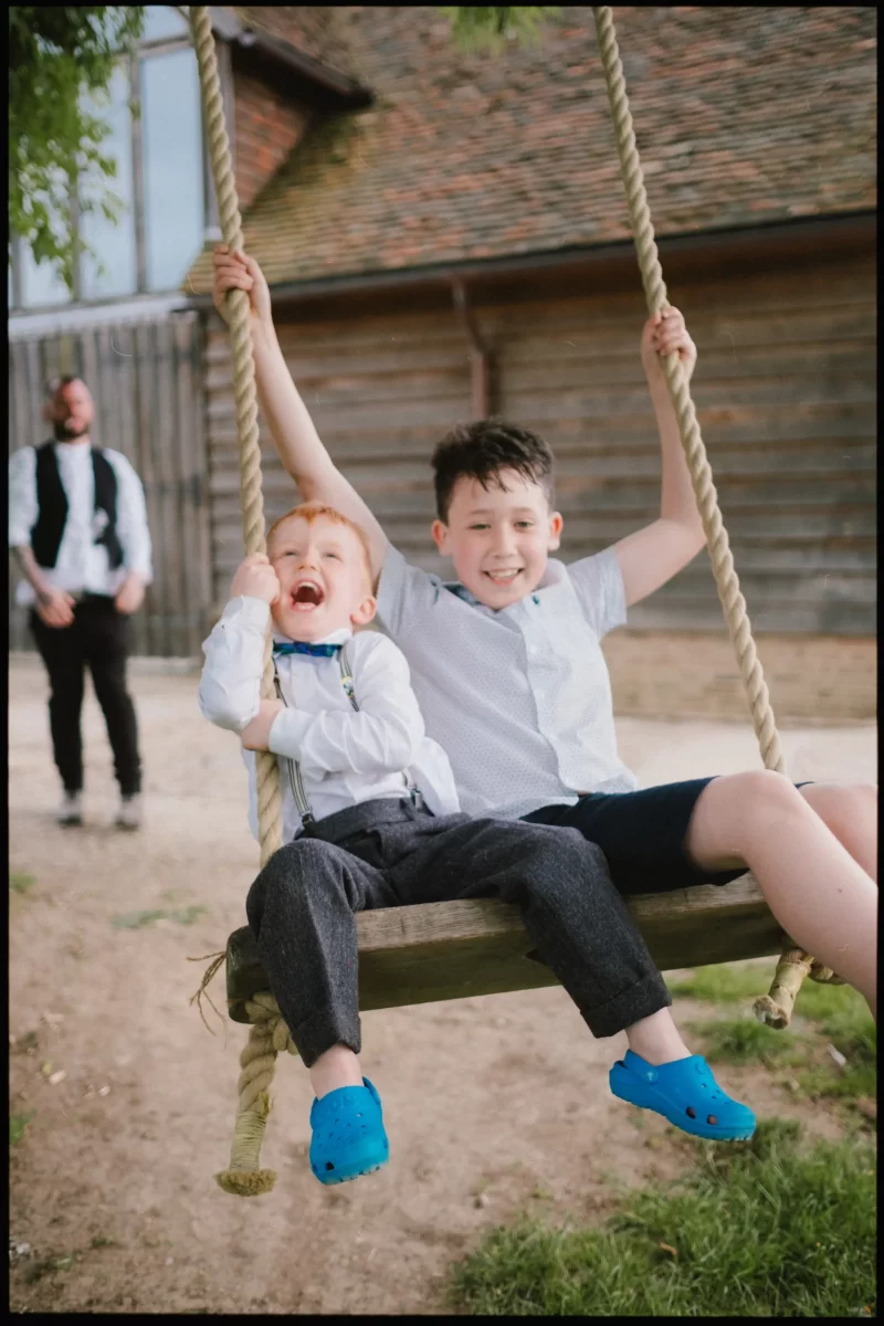 Two young boys sit together on a rope swing, smiling and laughing at a yoghurt rooms wedding festival. One wears a bow tie and suspenders; the other shorts and blue sandals. An adult stands in the blurred background near a wooden building.