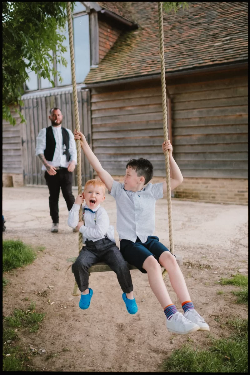 Two young boys sit together on a rope swing, smiling and playing at a Yoghurt Rooms wedding festival. One boy wears grey pants and blue shoes, the other wears shorts and trainers. A man stands in the background near a wooden building.