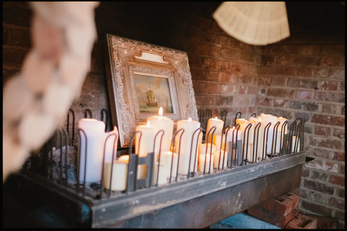 A metal tray holds numerous lit candles in front of a framed painting of a ship, all set against a rustic brick wall—evoking the inviting charm of yoghurt rooms wedding festival celebrations.