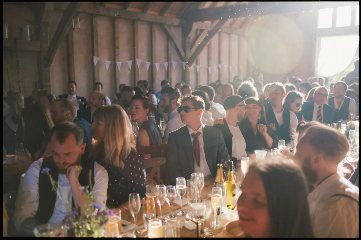 A large group of people sit at long tables decorated with glasses and bottles, enjoying a yoghurt rooms wedding festival in a rustic, sunlit barn with wooden beams and bunting. Some guests are dressed formally and smiling.