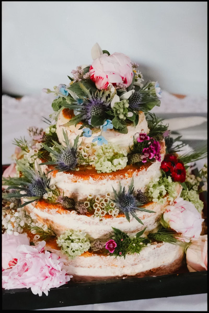 A three-tiered naked cake, perfect for a yoghurt rooms wedding festival, is adorned with fresh, colorful flowers and greenery—pink peonies, blue thistles, and small white blossoms—beautifully displayed on a black tray.