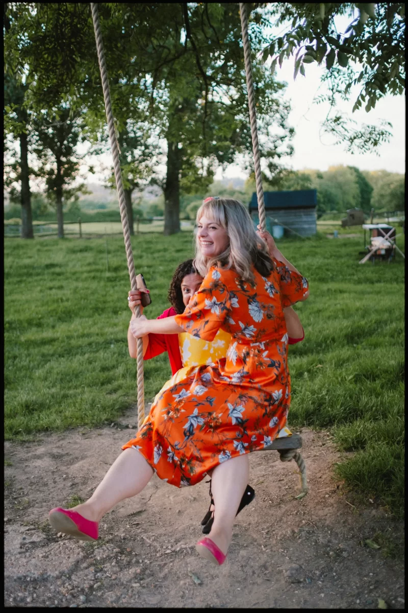 A woman in an orange floral dress and a child in a red shirt smile as they sit together on a rope swing outdoors, enjoying the sunny day at a yoghurt rooms wedding festival surrounded by green grass and trees.