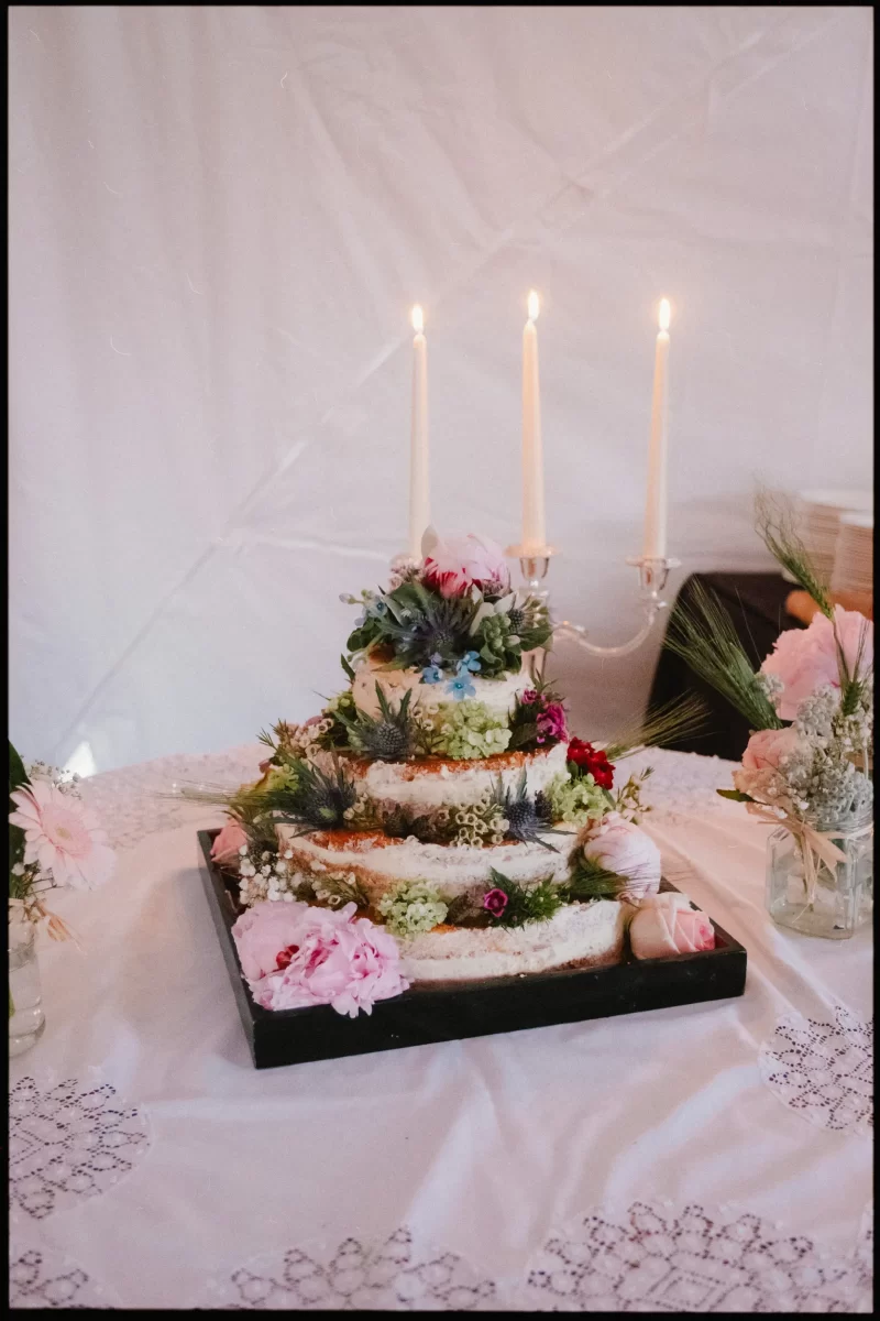 A three-tiered naked cake decorates this yoghurt rooms wedding festival scene, adorned with flowers and greenery, topped with white candles on a black tray atop a lace-covered table surrounded by small floral arrangements.