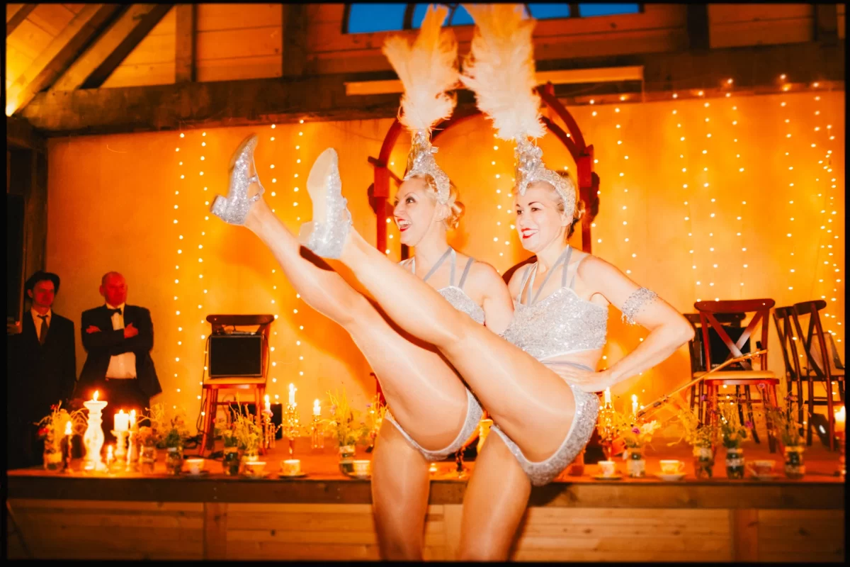 Two showgirls in sparkling silver costumes and feathered headpieces perform a high-kick dance routine on stage at a yoghurt rooms wedding festival. Warm orange lighting and string lights illuminate the festive scene.