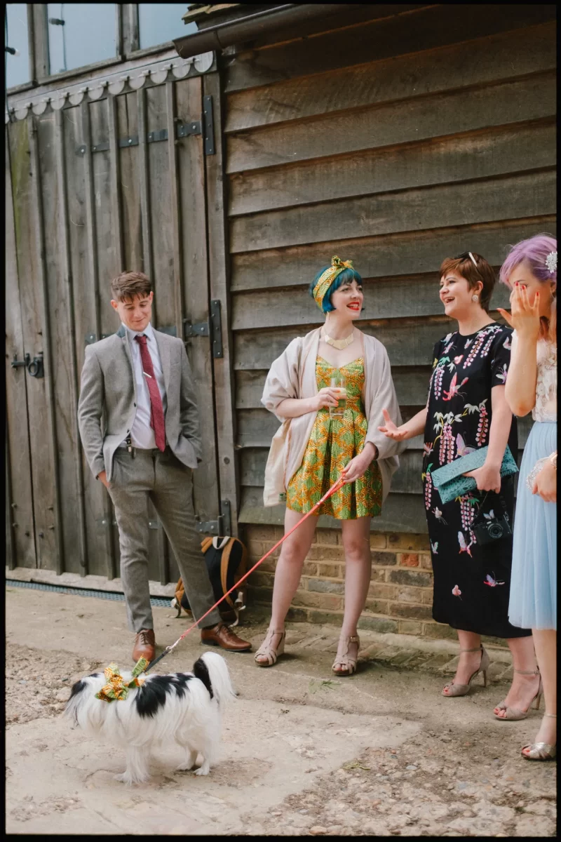 Four people in semi-formal, colorful clothes chat by a wooden building at a Yoghurt Rooms wedding festival. In front of them, a small white dog on a red leash, sporting a flower collar, completes the lively scene.