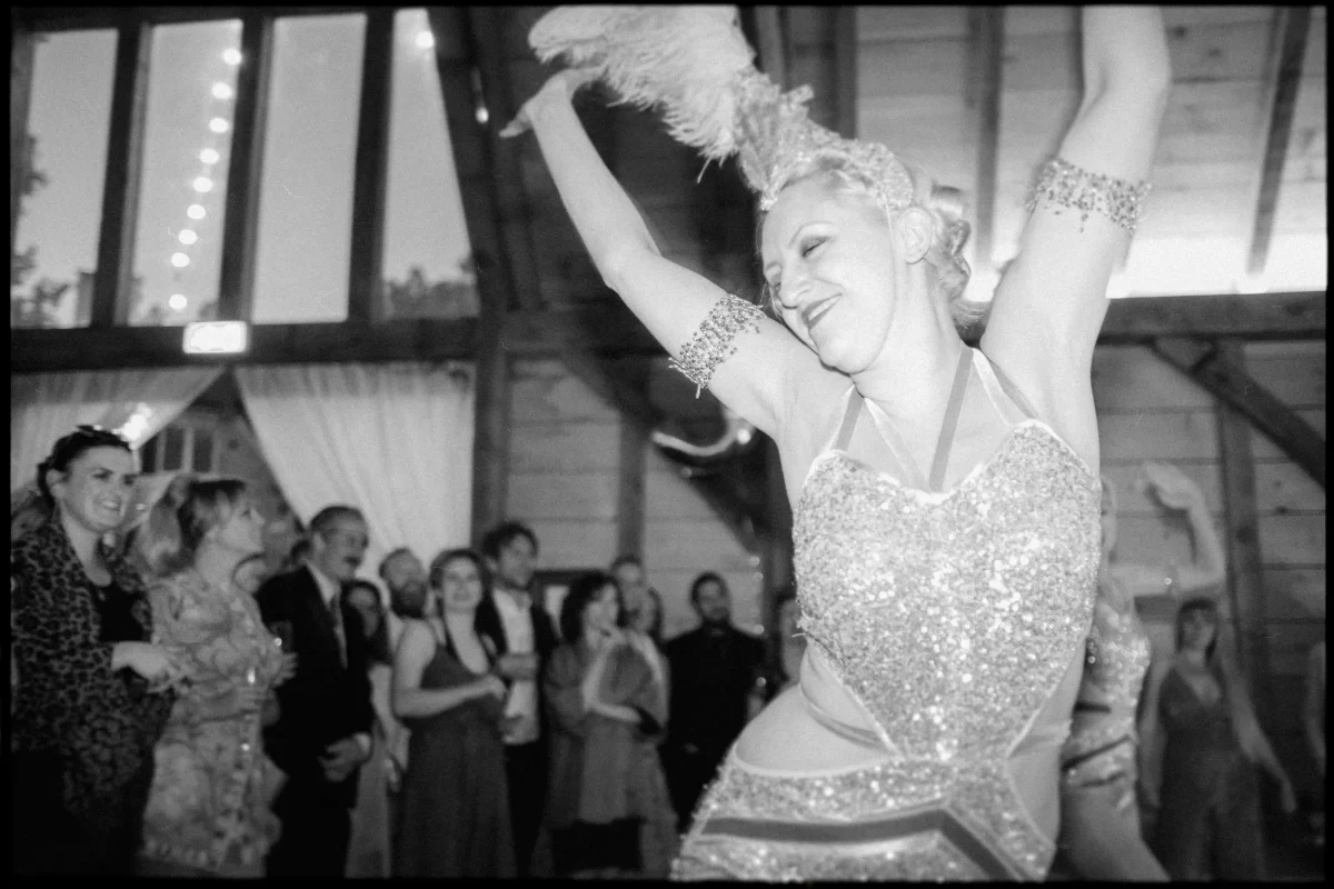 A performer in a sparkling costume and feathered headdress dances energetically at a yoghurt rooms wedding festival, delighting the smiling, applauding audience inside the rustic, wooden venue.