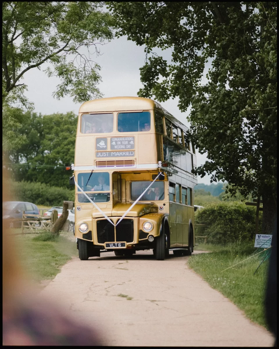 A vintage yellow double-decker bus, decorated with white ribbons and a “Just Married!” sign, drives down a country lane surrounded by trees—perfect for a Yoghurt Rooms wedding festival celebration.