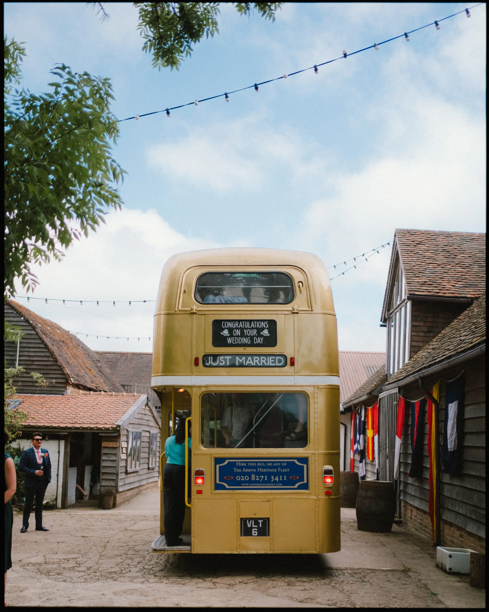 A vintage yellow double-decker bus with JUST MARRIED and wedding congratulations signs is parked between rustic wooden buildings at a sunny Yoghurt Rooms wedding festival, under string lights. People stand nearby.