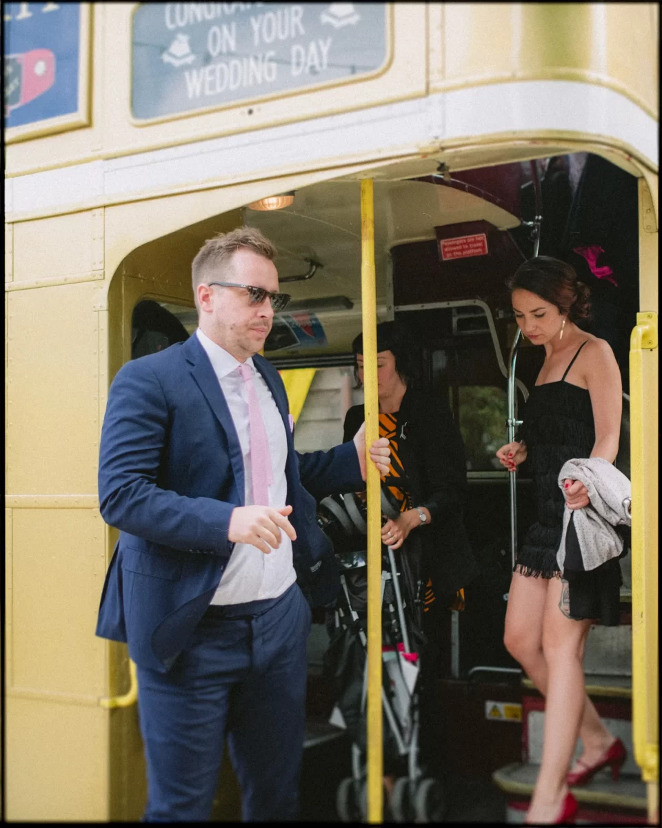 A man in a suit and sunglasses steps off a vintage yellow bus at the Yoghurt Rooms wedding festival, while a woman in a black dress and red heels follows behind him, holding her shoes and a jacket.