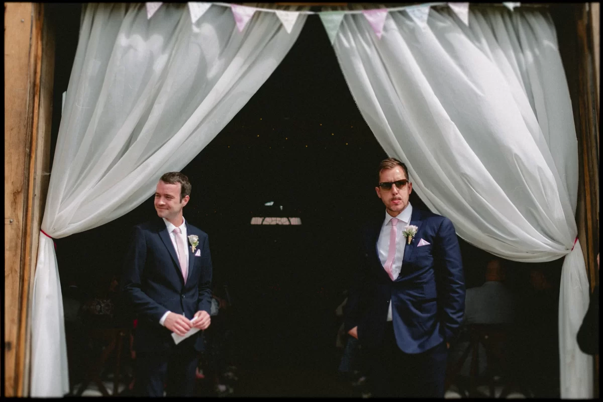 Two men in navy suits with pink ties stand before white curtains at an entrance, possibly at a yoghurt rooms wedding festival. One wears sunglasses; both have boutonnieres, suggesting a formal celebration is underway.