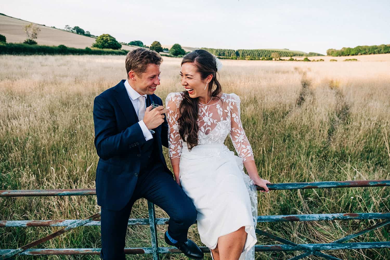 A bride and groom sit on a metal gate in a grassy field at Upwaltham Barns, laughing together; the groom wears a navy suit, and the bride wears a white dress with lace sleeves, with hills and trees in the background.