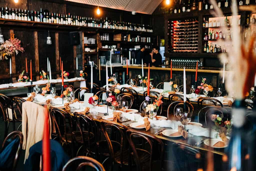 A long dining table set for an event in a dimly lit restaurant, decorated with flowers, candles, glassware, and plates. Wooden chairs surround the table, at sager and wildel. Shelves of bottles line the back wall.
