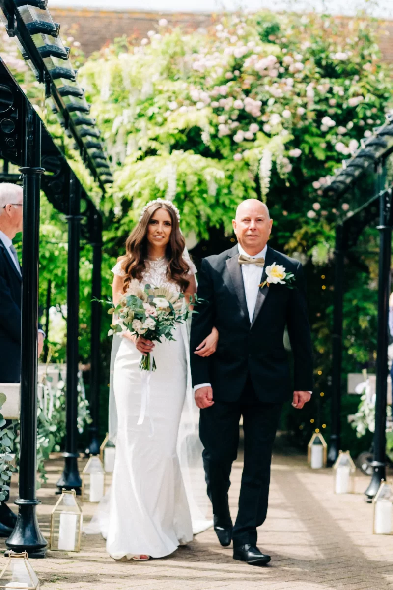 A bride in a white dress holding a bouquet walks arm-in-arm with a man in a black suit and yellow boutonniere down an outdoor aisle at an Old Kent Barn wedding in Dover, Kent, decorated with greenery and lanterns.