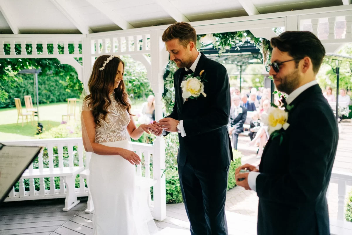 A bride and groom stand in a gazebo during their Old Kent Barn wedding in Dover, Kent. The groom places a ring on the bride’s finger as the best man stands beside them holding a ring box, with guests seated outside in the background.