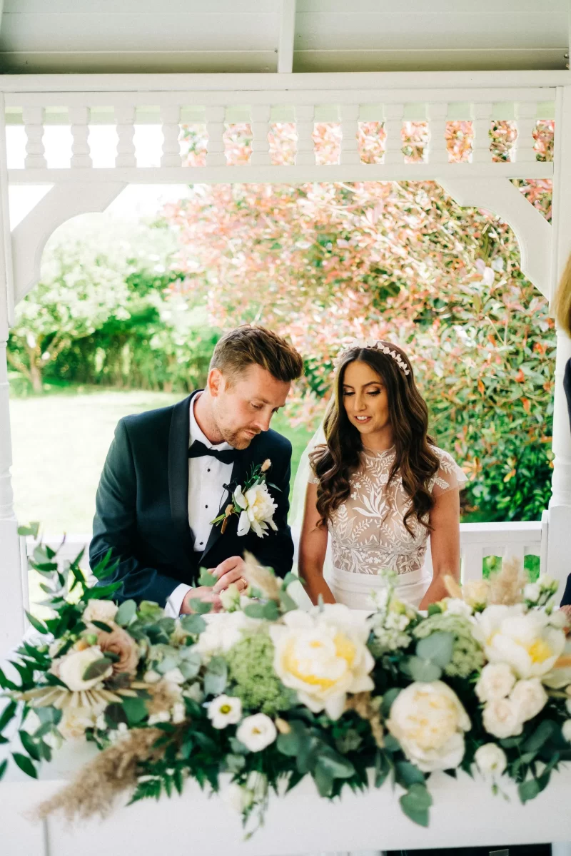 A bride and groom sit side by side at a white gazebo table decorated with lush white and green flowers at their Old Kent Barn wedding in Dover, Kent. The groom wears a black tuxedo, the bride wears a lace dress with loose curls and a pearl headpiece.