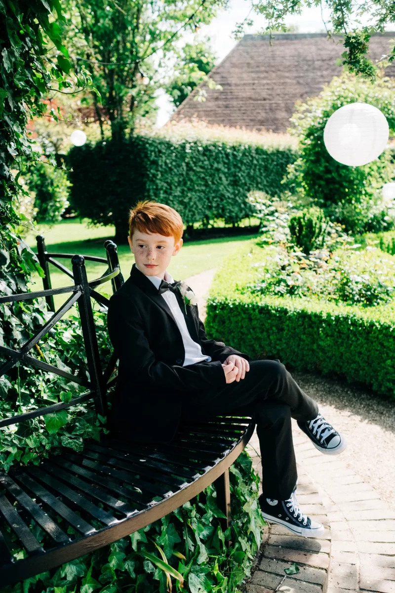A red-haired boy in a black suit and bow tie sits on a black metal bench in a lush garden at an Old Kent Barn wedding near Dover, Kent. He glances to the side, wearing black sneakers amid green hedges and white paper lanterns.