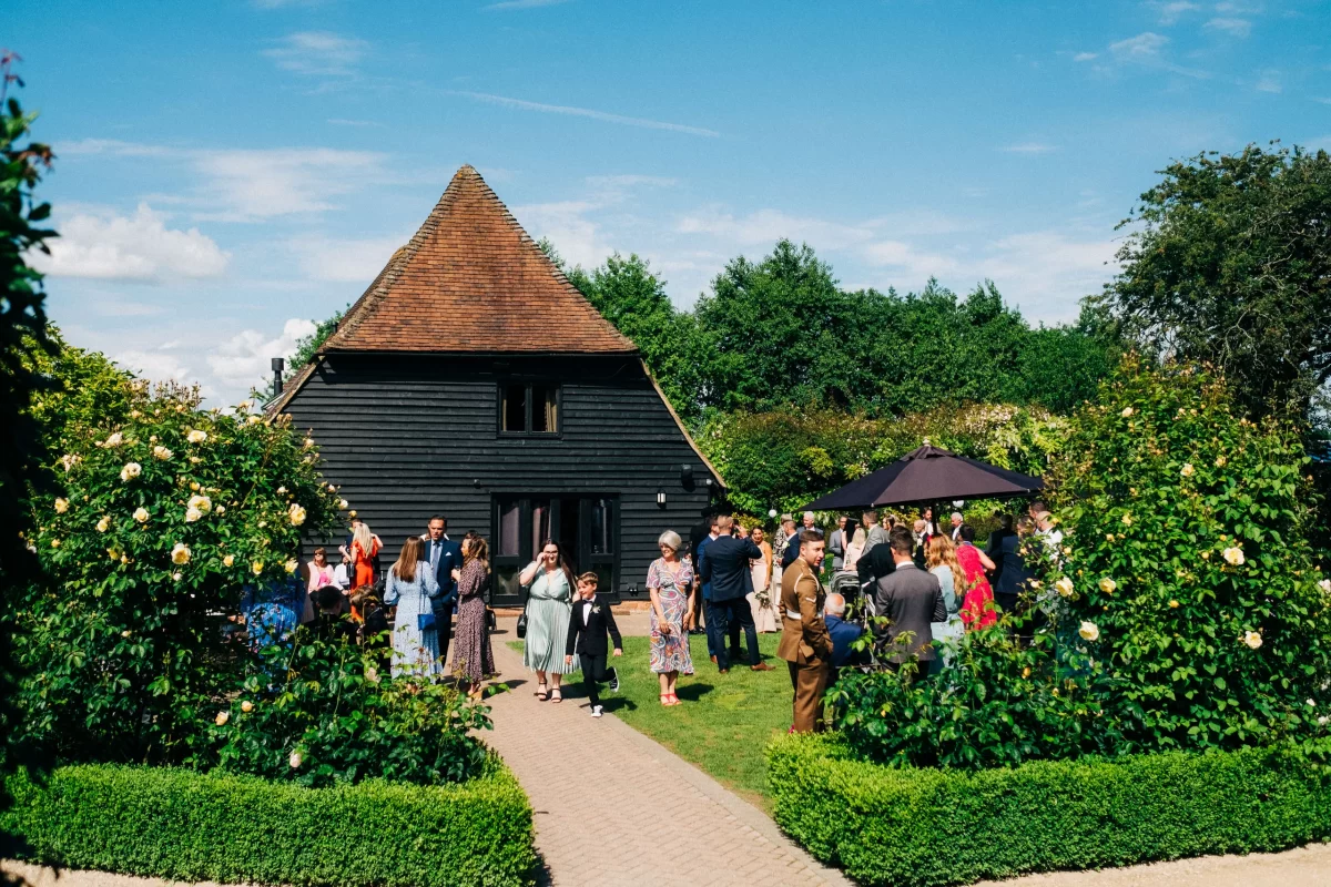 A group of people dressed in formal attire gather outside the Old Kent Barn, a black timber building with a steep roof, surrounded by green bushes and flowers under a bright blue sky—perfect for a wedding in Dover, Kent.