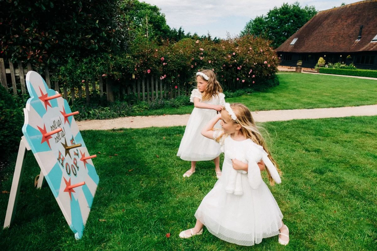 Two young girls in white dresses play a throwing game with red beanbags at an outdoor event on green grass, with flowers, a house, and the charming Old Kent Barn wedding venue near Dover, Kent in the background.