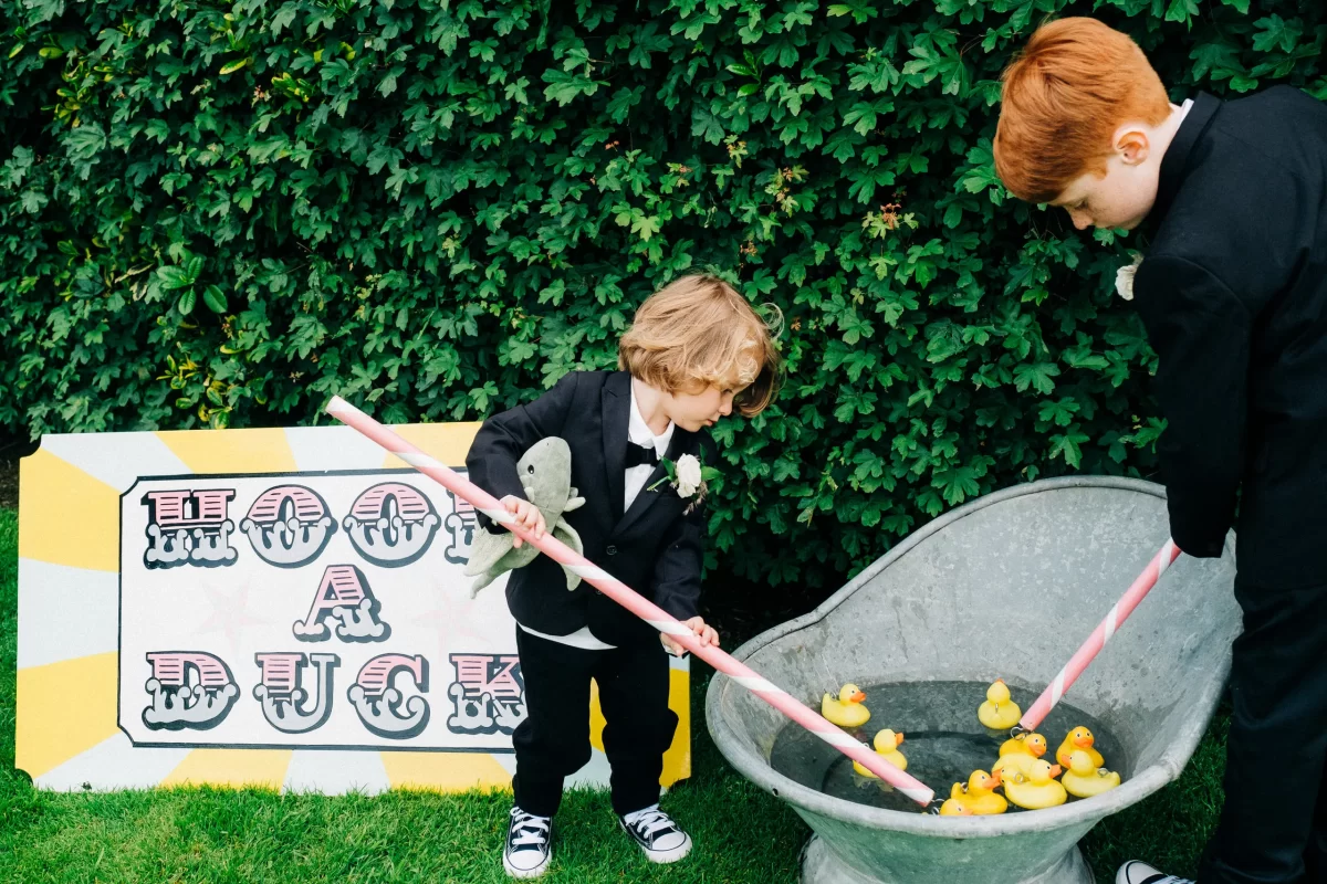 Two young boys in suits play hook a duck outdoors at an Old Kent Barn wedding near Dover, Kent, using long sticks to fish rubber ducks from a tub. A colorful "Hook a Duck" sign stands out against lush green bushes behind them.