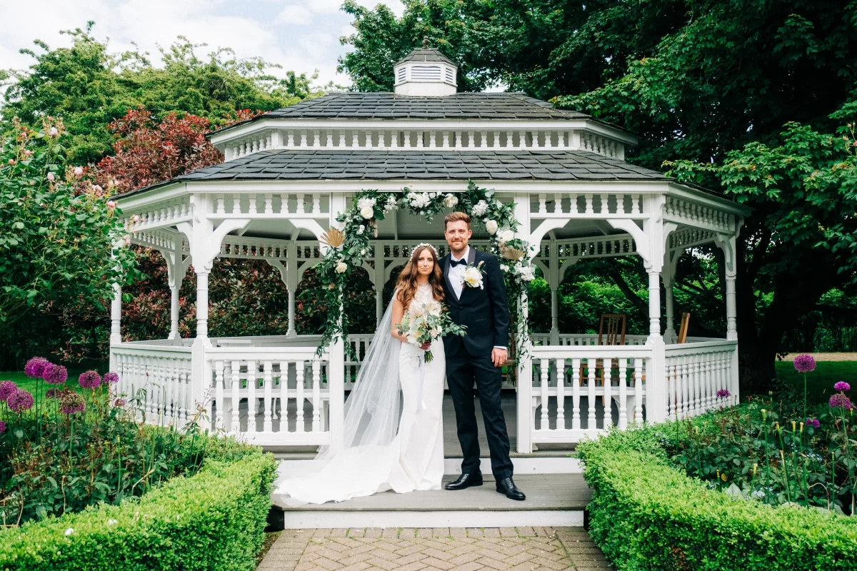 A bride in a white dress and veil stands next to a groom in a black suit and bow tie before a flower-adorned gazebo at an Old Kent Barn wedding near Dover, Kent, surrounded by lush greenery and blooming gardens.