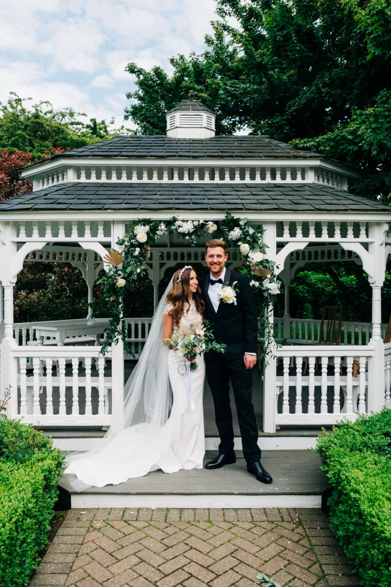 A bride in a white dress and veil stands next to a groom in a black tuxedo, both smiling, in front of a white gazebo decorated with greenery and white flowers at an Old Kent Barn wedding near Dover, Kent.