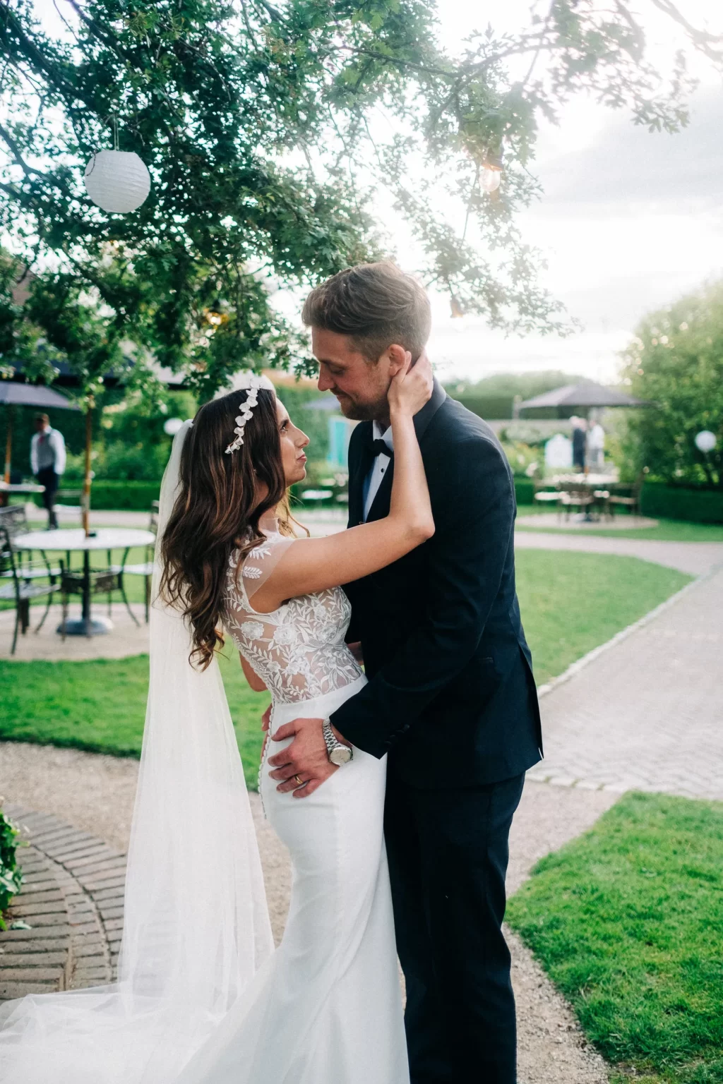 A bride and groom embrace outdoors on a lush green lawn at an Old Kent Barn wedding in Dover, Kent. The bride wears a white dress and veil, while the groom is in a black suit. They gaze at each other lovingly under lanterns and leafy trees.