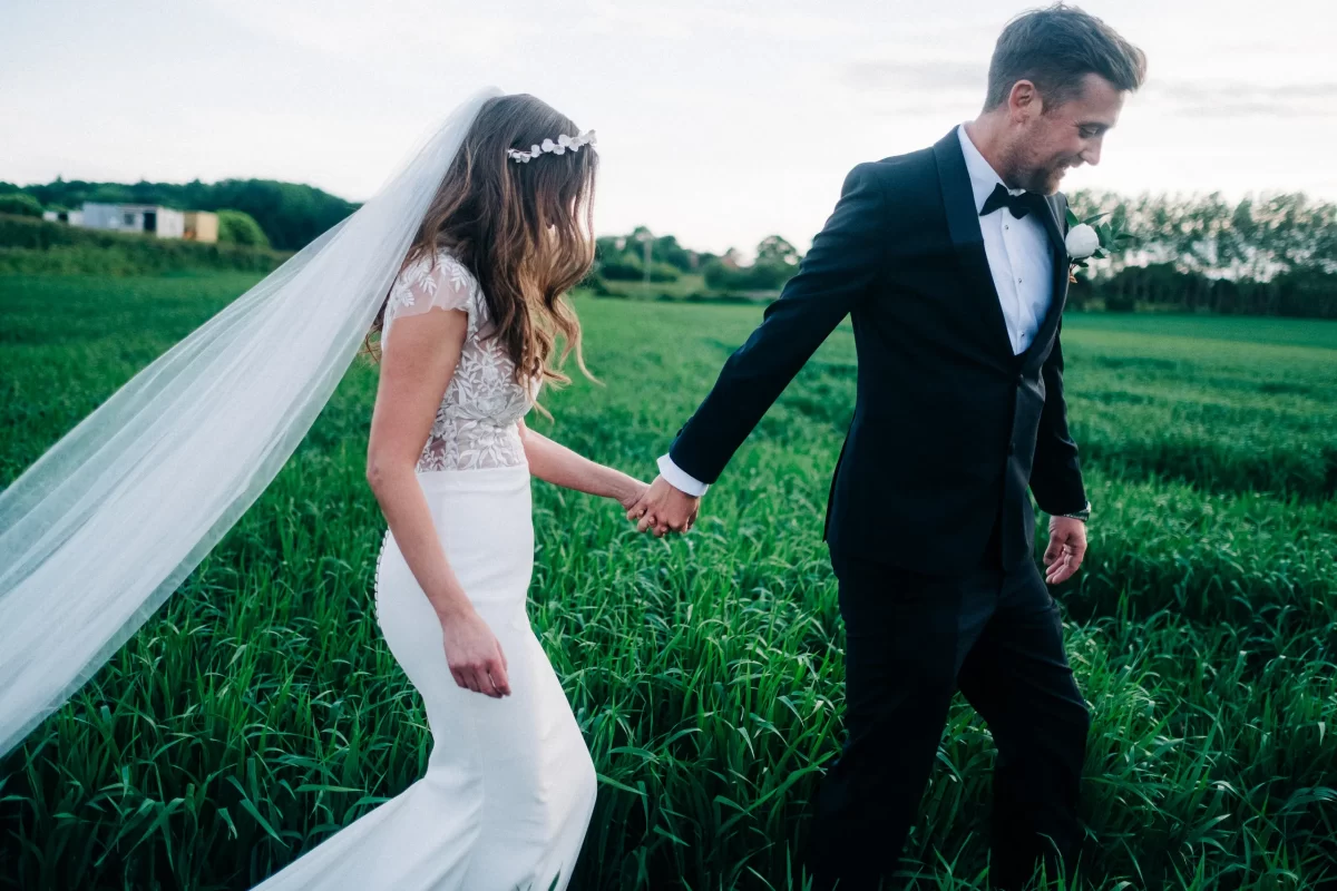 A bride in a white dress and veil holds hands with a groom in a black tuxedo as they walk through a green field at Old Kent Barn, Dover Kent, smiling and looking at each other.