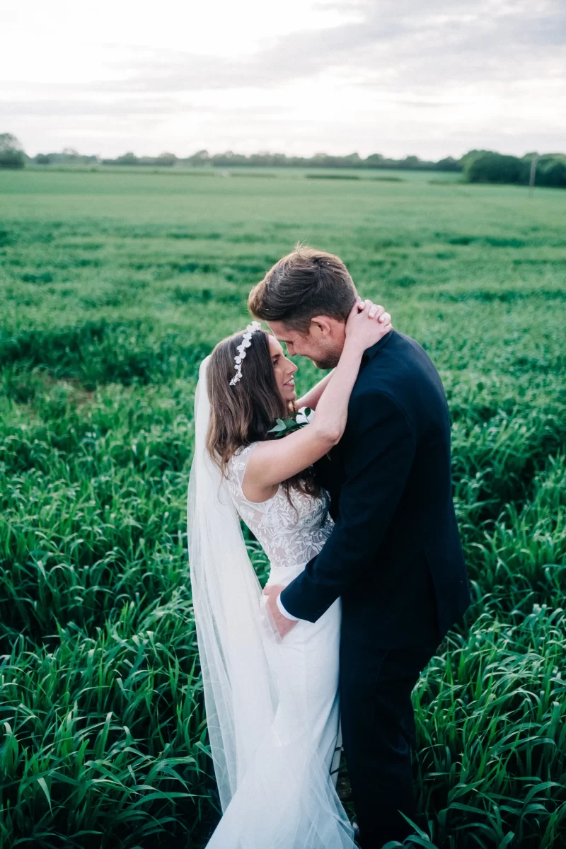 A bride and groom embrace and smile at each other in a lush green field at Old Kent Barn, with the bride in a white dress and veil and the groom in a black suit, under a partly cloudy sky—capturing the magic of a wedding in Dover, Kent.