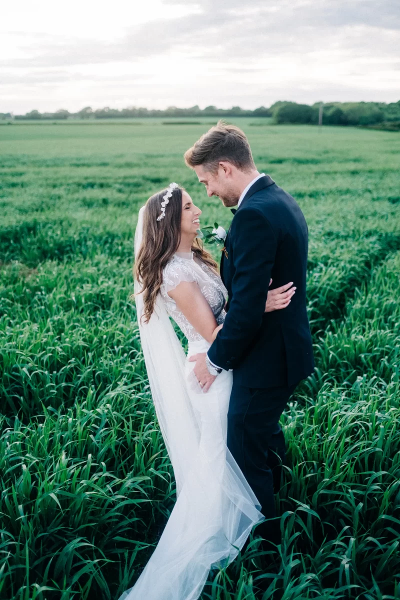 A bride and groom stand in a green field at Old Kent Barn, Dover, Kent, gazing and smiling at each other. The bride wears a white dress with a veil and headpiece, while the groom is in a dark suit against open farmland under a cloudy sky.