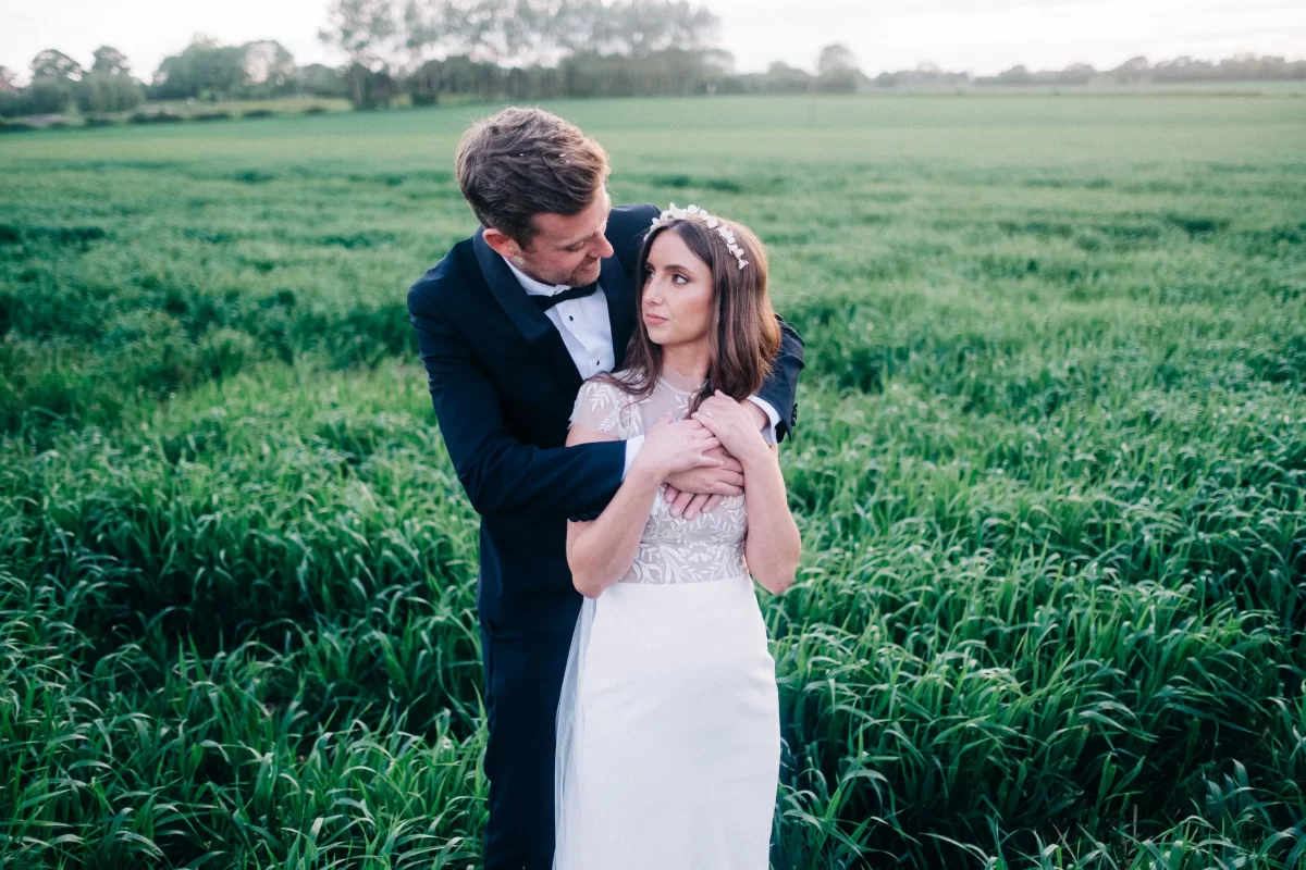 A groom in a black suit embraces a bride in a white dress and floral headpiece as they stand in a lush green field at Old Kent Barn, Dover, Kent, gazing at each other with trees and sky in the background.