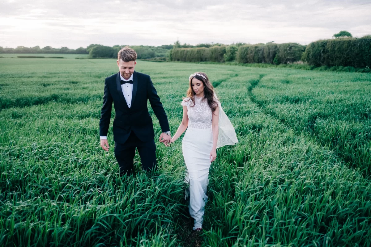 A bride and groom walk hand in hand through a green field at the Old Kent Barn. The groom wears a black tuxedo and bow tie; the bride, a white dress with veil and jeweled headpiece. The lush Dover, Kent setting is beautifully outdoors.