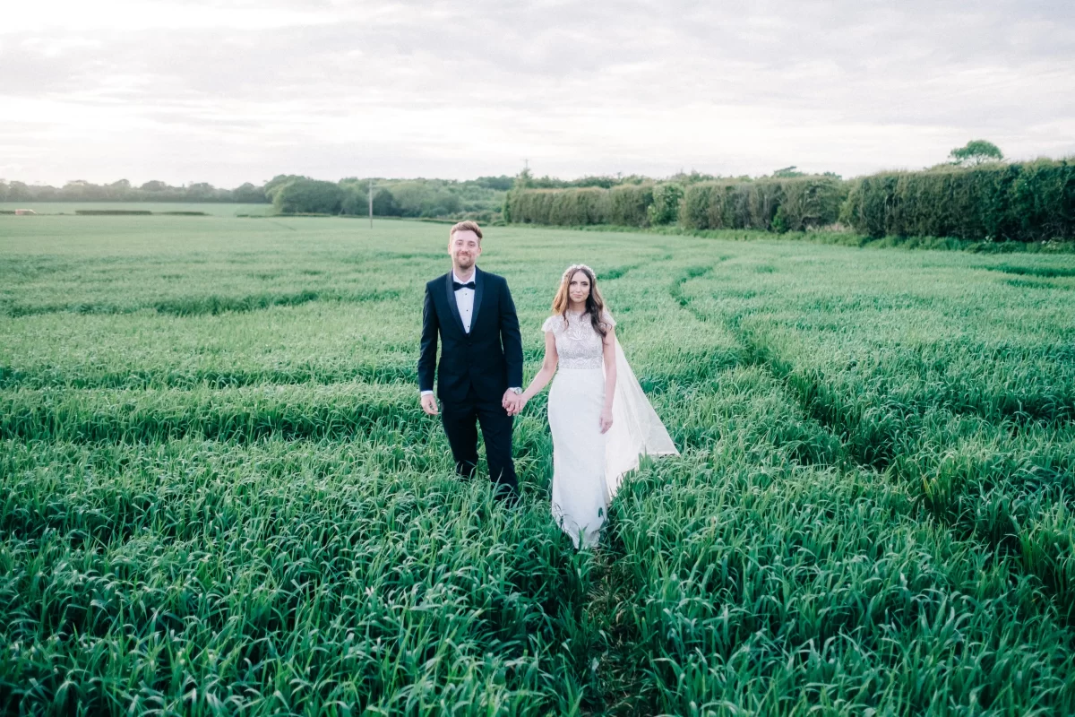 A bride and groom walk hand in hand through a green field on a cloudy day at an Old Kent Barn wedding in Dover, Kent. The bride wears a white dress and veil, while the groom is in a dark suit with a bow tie. Lush greenery stretches out behind them.