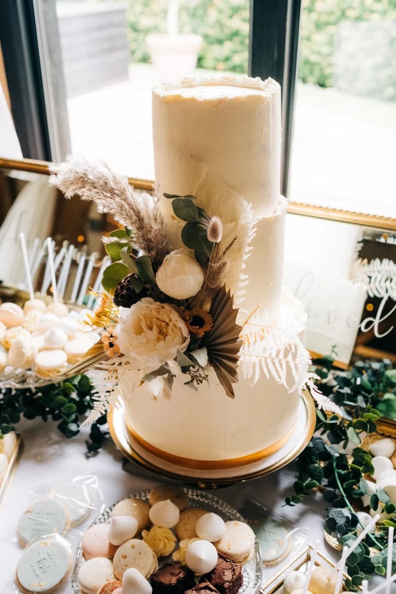 A tall, white, two-tiered wedding cake decorated with white and beige flowers, greenery, and dried foliage sits on a gold-rimmed stand with assorted macarons—perfect for an Old Kent Barn wedding in Dover, Kent.