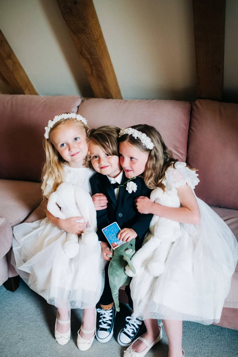 Three young children sit on a pink sofa at an Old Kent Barn wedding in Dover, Kent. Two girls in white dresses and flower headbands hug a boy in a black suit and bow tie. The girls hold stuffed bunnies; the boy holds a toy dinosaur and blue card.