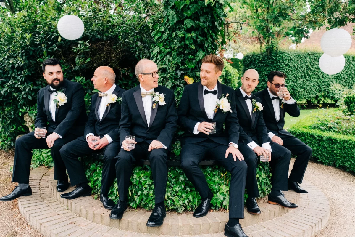 Six men in black tuxedos with boutonnieres sit together on a circular brick bench in a lush garden at an Old Kent Barn wedding in Dover, Kent, holding drinks and chatting, surrounded by greenery and white paper lanterns.