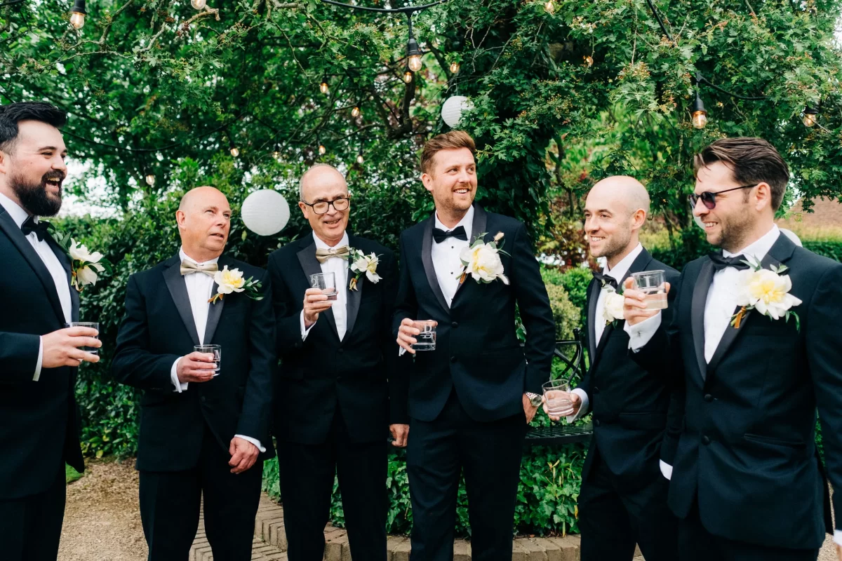 Six men in black tuxedos with white boutonnieres stand outdoors, smiling and holding drinks beneath green trees with string lights and white paper lanterns, celebrating at an Old Kent Barn wedding in Dover, Kent.
