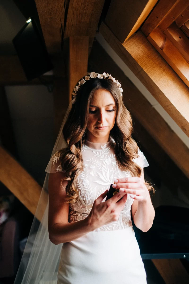 A bride with long wavy hair and a flower crown stands indoors by a window at an Old Kent Barn wedding in Dover, Kent, wearing a white lace dress and holding a small object in her hands, looking down thoughtfully.