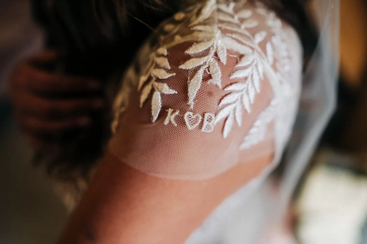 A close-up of a bride’s shoulder at an Old Kent Barn wedding in Dover, Kent, shows white lace embroidery with leafy patterns and the initials K ♡ B stitched onto sheer fabric.