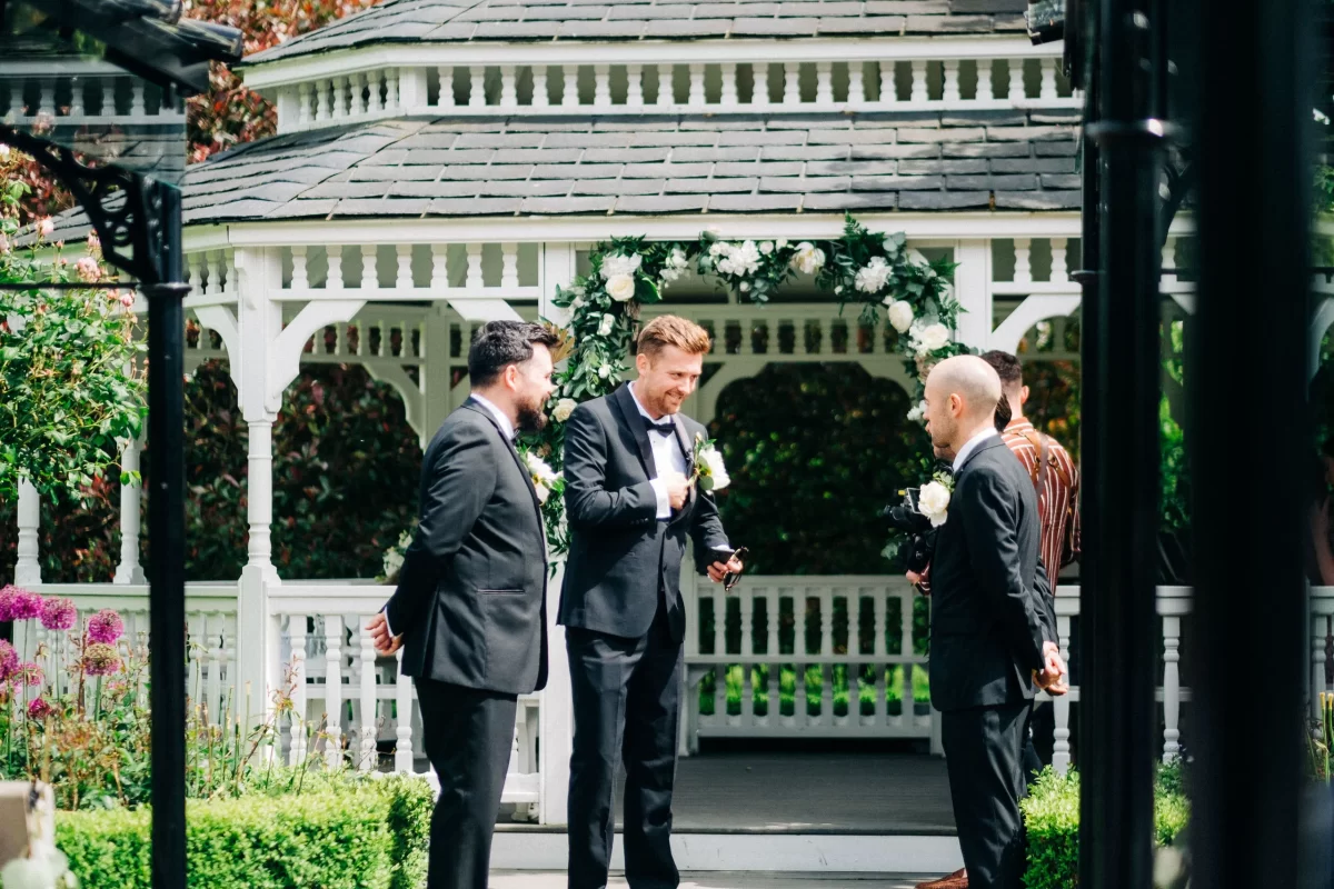 Three men in suits chat in front of a white gazebo adorned with greenery and flowers at an Old Kent Barn wedding, enjoying the sunny garden setting near Dover, Kent.