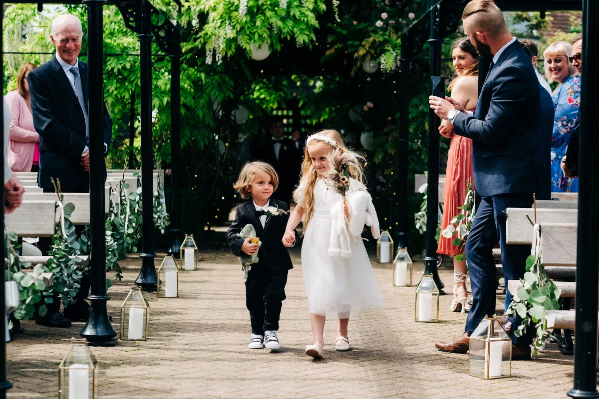 A young boy in a black suit and a girl in a white dress walk down the outdoor aisle at an Old Kent Barn wedding near Dover, Kent, surrounded by smiling guests and greenery, with lanterns lining the path.