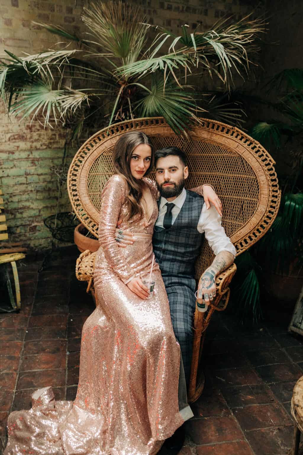 bride and groom posing on retro chair bride in pink sequin wedding dress, groom relaxed with sleeves up, bride sitting on his lap