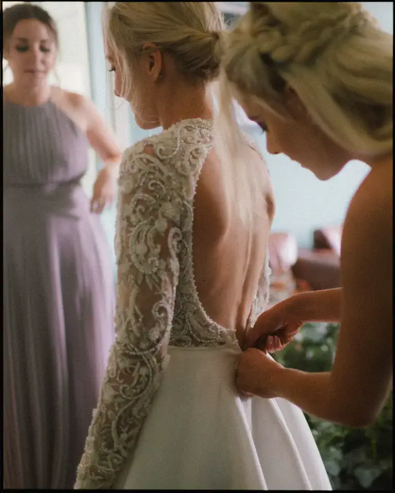 A bride in a lace, open-back wedding dress has her gown adjusted by a bridesmaid while another woman in a lavender dress stands nearby, all beautifully captured by a documentary London wedding photographer in softly lit, candid style.